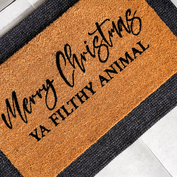 Natural coir doormat with 'Merry Christmas' in elegant script above 'Ya Filthy Animal' in bold block letters, photographed [a